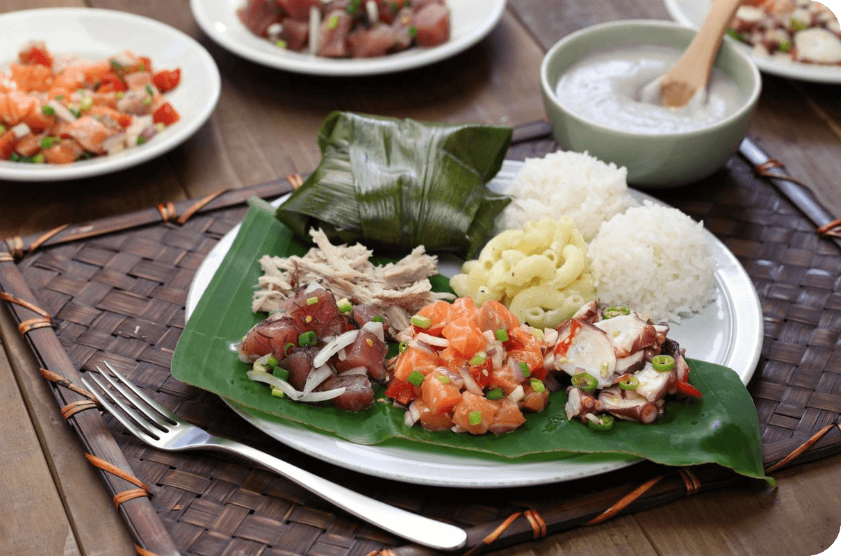 A traditional hawaiian meal featuring poke, rice, and side dishes served on a banana leaf and a woven mat.