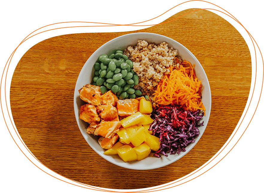 A colorful bowl of food with sections of grilled chicken, quinoa, edamame, shredded carrots, mango, and purple cabbage on a wooden table.